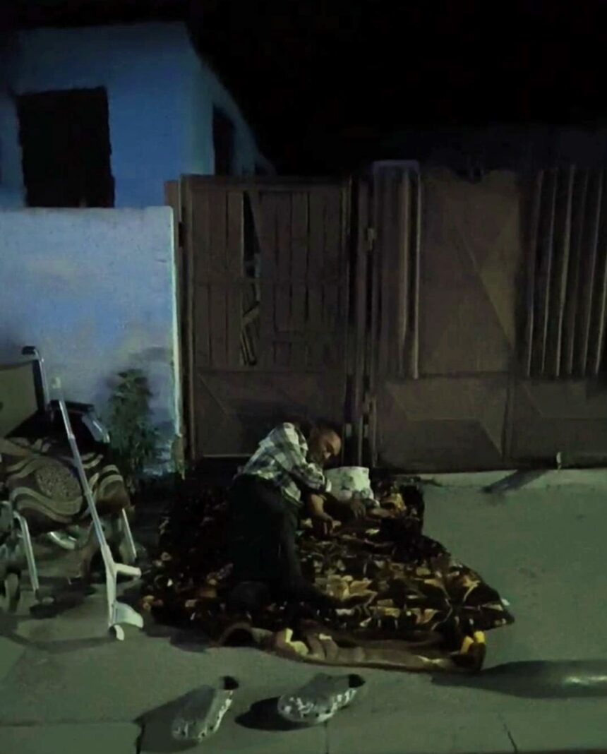 Man kneeling on a patterned blanket at night near a wooden gate, with shoes and a chair nearby in a dim outdoor area.