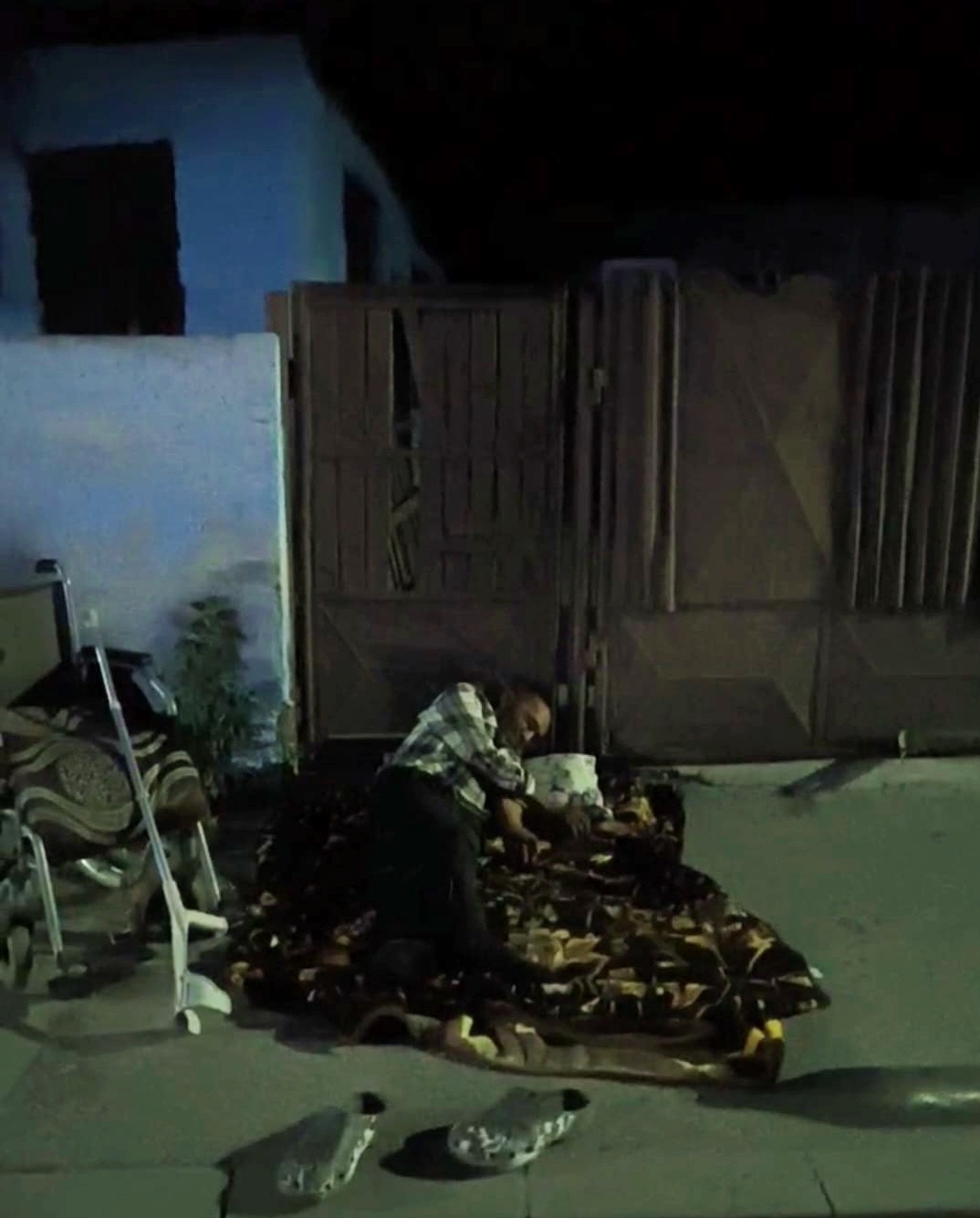 Man kneeling on a patterned blanket at night near a wooden gate, with shoes and a chair nearby in a dim outdoor area.