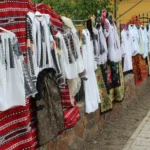 Row of traditional white shirts with black embroidery and red fabrics hung on a fence at an outdoor market.