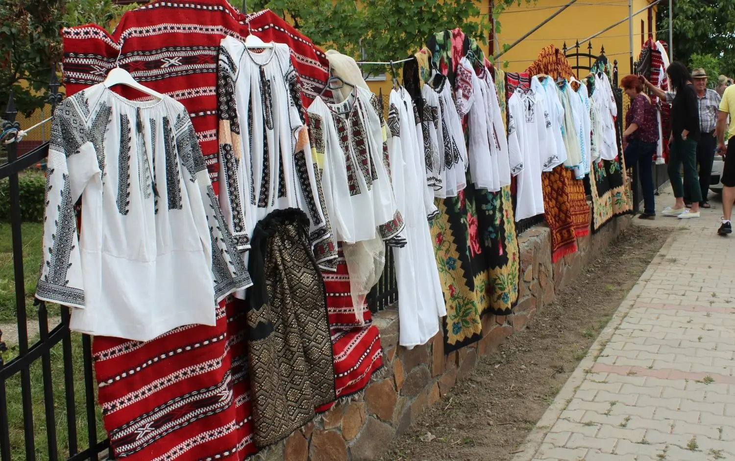 Row of traditional white shirts with black embroidery and red fabrics hung on a fence at an outdoor market.