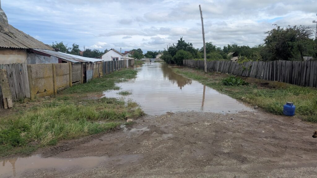 Flooded rural street with standing water along a dirt road, fences and modest houses on both sides; a blue barrel on the right.