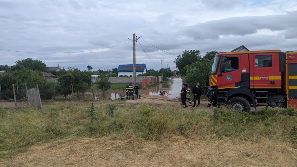 Red fire truck on a muddy street with firefighters near a flooded road in a rural area, utility poles and houses in the background