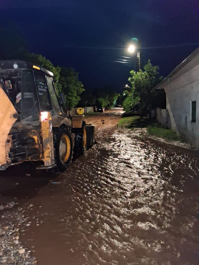 Nighttime street scene with a large construction vehicle on the left beside a muddy, reflective road and trees lining the street.