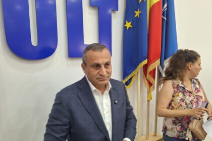 Two adults seated indoors at a desk, with blue flags featuring yellow stars behind them.