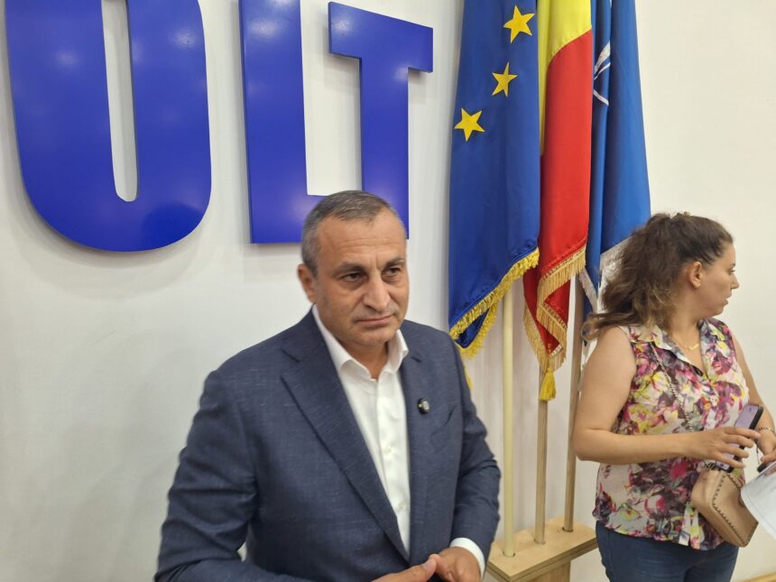 Two adults seated indoors at a desk, with blue flags featuring yellow stars behind them.