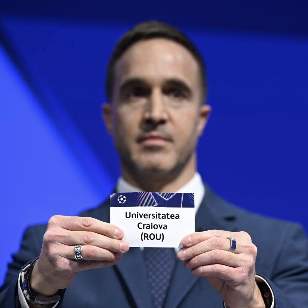 Man in a suit holds a card reading Universitatea Craiova (ROU) against a blue background.