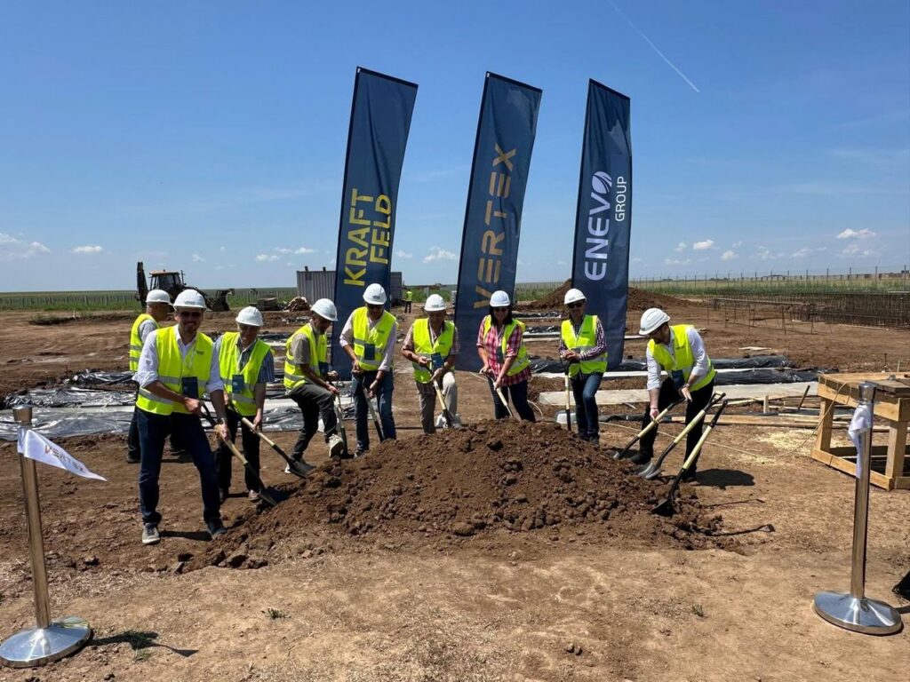 Group of people in hard hats and high-visibility vests break ground with shovels at a construction site, flags in the background under a clear sky.