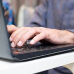 Close-up of hands typing on a laptop keyboard on a white desk nearby.