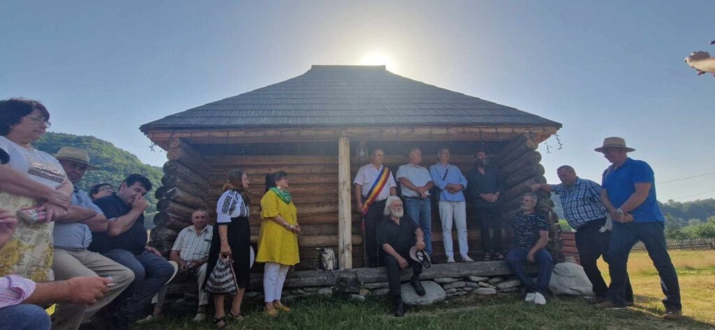 Group of people gathered on a rustic log cabin porch, sun behind the roof, likely a community event or ceremony.