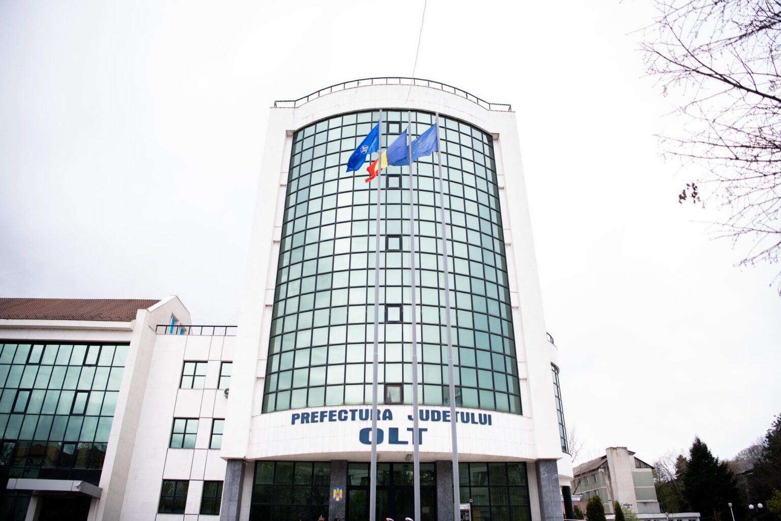 Front view of a white office building with a curved glass facade and three flagpoles in front, flags fluttering outside.