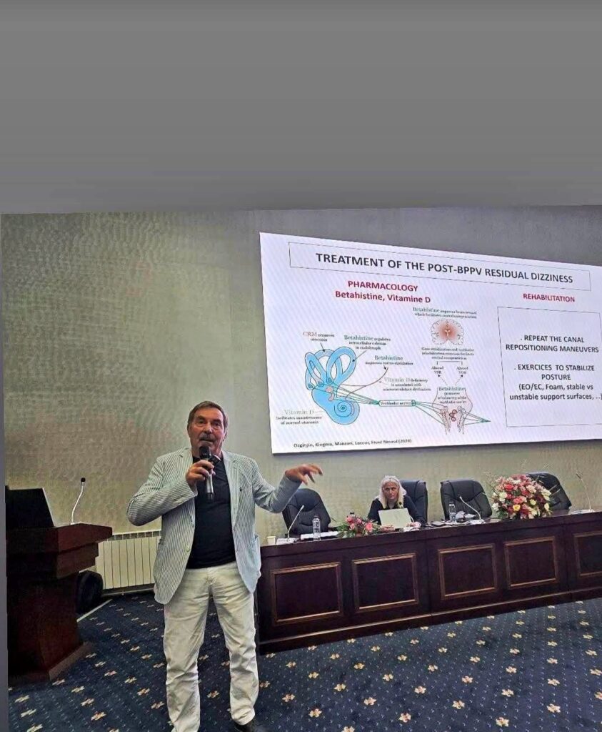 prof laquer - Jurnalul Olteniei – Știri din Craiova și Oltenia Man in a light striped blazer speaks into a handheld microphone at a conference, gesturing toward a large medical poster on post-BPPV dizziness behind him.