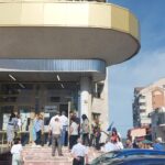 Crowd of people lined up outside a modern building with glass doors and a curved yellow balcony on a sunny day.