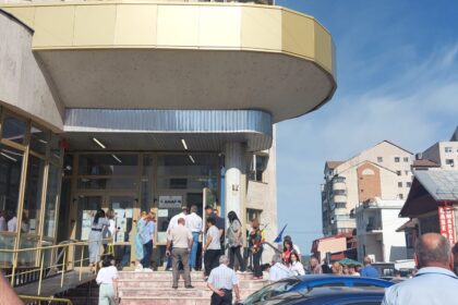 Crowd of people lined up outside a modern building with glass doors and a curved yellow balcony on a sunny day.