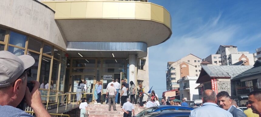 Crowd of people lined up outside a modern building with glass doors and a curved yellow balcony on a sunny day.