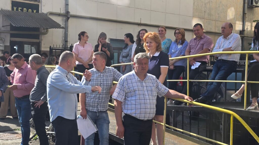 People gather on a yellow metal ramp outside a beige building, some chatting and watching an event unfold.