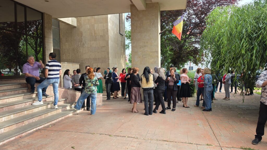 Group of people chatting outside a beige stone building near the entrance, with a flag on a pole visible overhead.