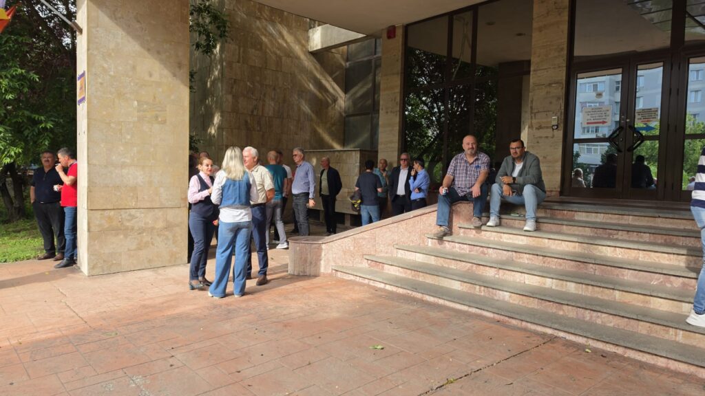Crowd of adults gathered outside a beige stone building with wide pink marble steps and glass entrance doors, some chatting.