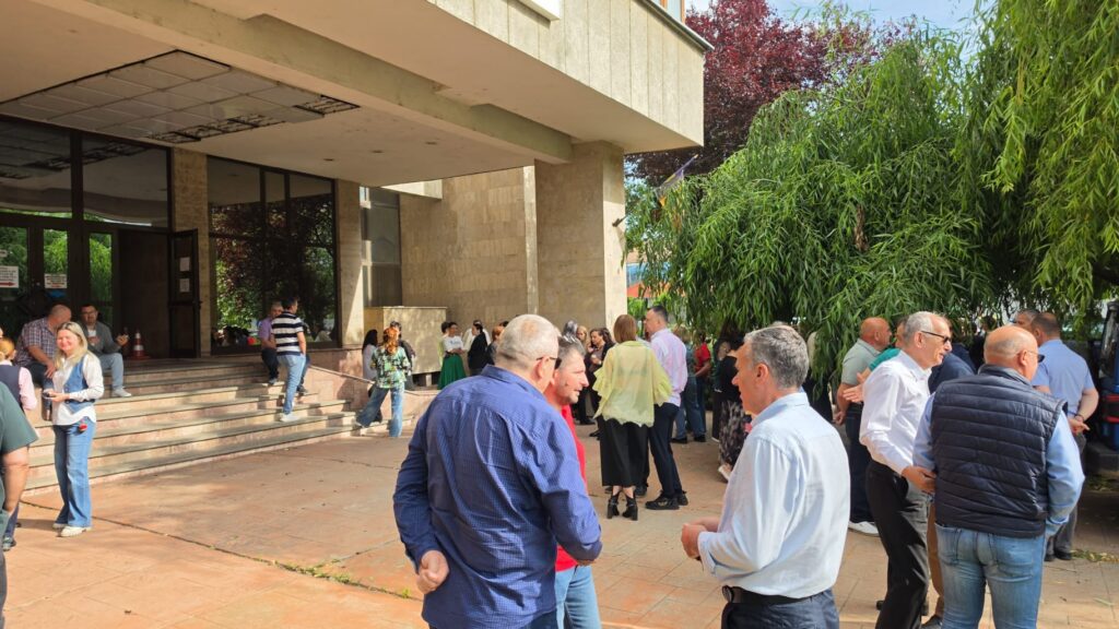 Crowd of adults gathered outside a beige stone building entrance, chatting near the stairs and glass doors.