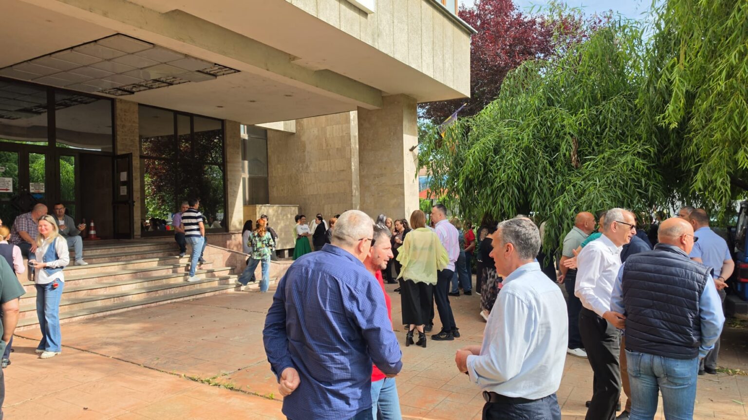 Crowd of adults gathered outside a beige stone building entrance, chatting near the stairs and glass doors.