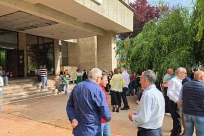 Crowd of adults gathered outside a beige stone building entrance, chatting near the stairs and glass doors.