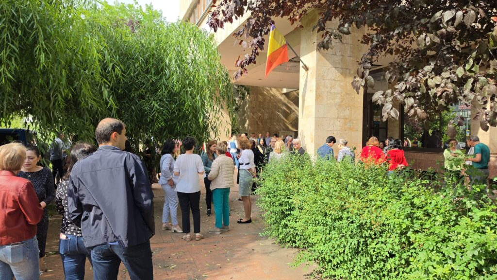 Group of people conversing outside a beige stone building with a colorful flag overhead, surrounded by green hedges and trees.