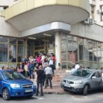 Crowd of people gathered outside a modern glass-fronted building entrance with Romanian flags on the wall and several parked cars in front of the scene.