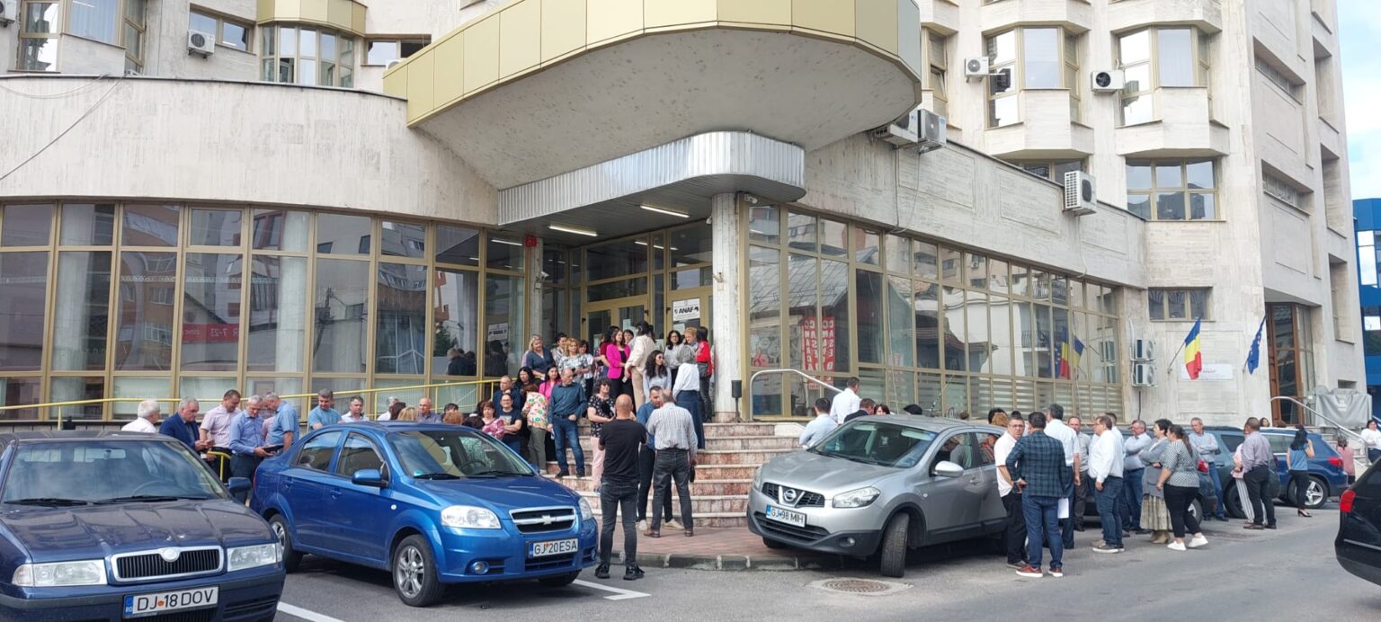 Crowd of people gathered outside a modern glass-fronted building entrance with Romanian flags on the wall and several parked cars in front of the scene.