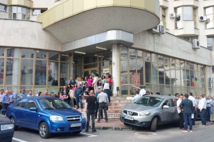 Crowd of people gathered outside a modern glass-fronted building entrance with Romanian flags on the wall and several parked cars in front of the scene.