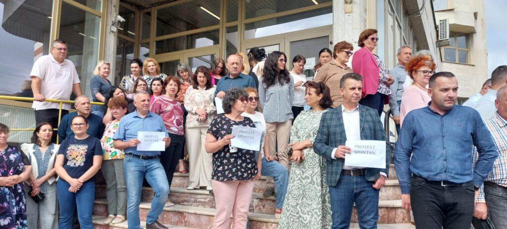 Group of people standing on steps outside a building, some holding protest signs that read ‘PROTEST SPONTAN’.