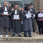 Group of people in dark legal robes stand on steps outside a stone building, holding signs