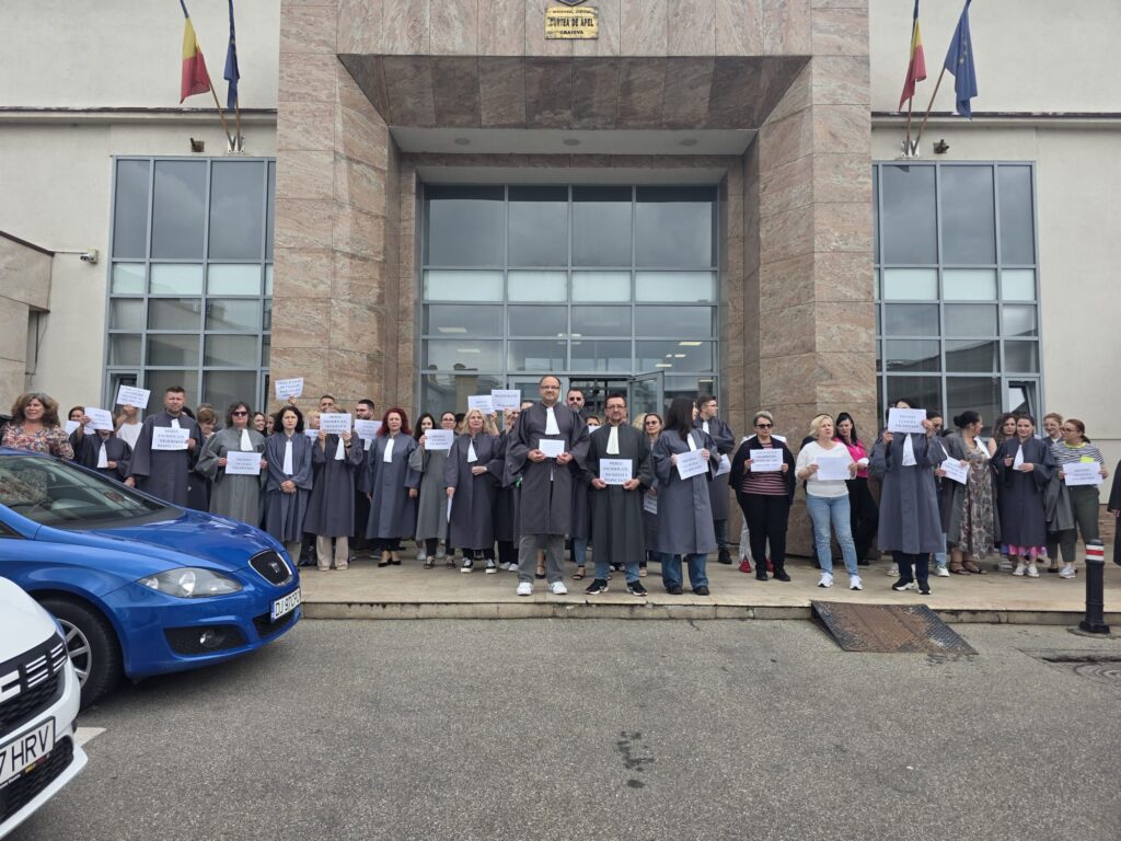 Row of graduates in gray robes standing outside a stone building, each holding a sign, with cars parked in the foreground and flags above the entrance.