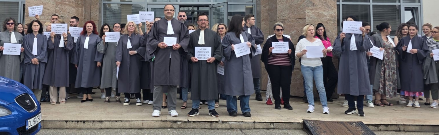 Group of people in dark legal robes stand on steps outside a stone building, holding signs