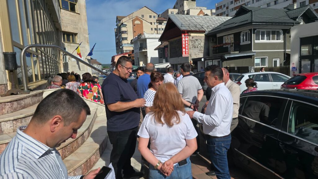 A group of people standing and chatting outside a building on a sunny day, with cars parked along the street.
