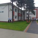 Exterior of a hospital building with the sign 'Investim în Sănătate'; several people in blue shirts stand near the entrance, benches, and a brick-paved path nearby.