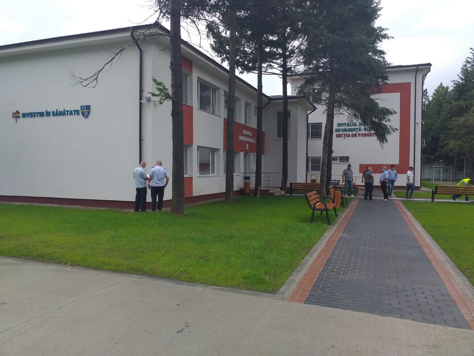 Exterior of a hospital building with the sign 'Investim în Sănătate'; several people in blue shirts stand near the entrance, benches, and a brick-paved path nearby.