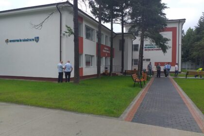 Exterior of a hospital building with the sign 'Investim în Sănătate'; several people in blue shirts stand near the entrance, benches, and a brick-paved path nearby.