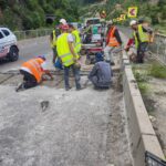 Construction workers in safety vests and helmets repair a mountain road with equipment and a concrete barrier on a windy hillside street
