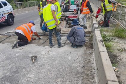 Construction workers in safety vests and helmets repair a mountain road with equipment and a concrete barrier on a windy hillside street