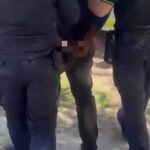 Two uniformed officers restrain a person standing between them in a street or outdoor area.