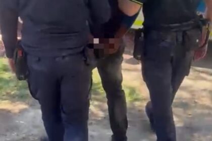 Two uniformed officers restrain a person standing between them in a street or outdoor area.