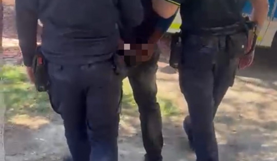 Two uniformed officers restrain a person standing between them in a street or outdoor area.