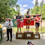 Three athletes stand on a wooden podium holding diplomas and flags (Portugal and Romania) in a park, with a bust statue and trees in the background.