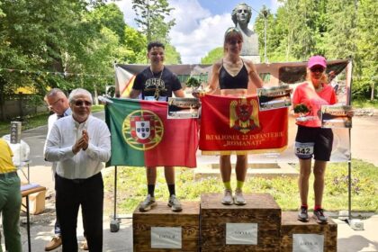Three athletes stand on a wooden podium holding diplomas and flags (Portugal and Romania) in a park, with a bust statue and trees in the background.