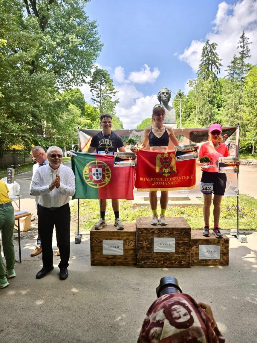 Three athletes stand on a wooden podium holding diplomas and flags (Portugal and Romania) in a park, with a bust statue and trees in the background.