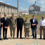 Group of military personnel and civilians posing for a photo outside a base on a sunny day, with uniformed and civilian attire side by side.