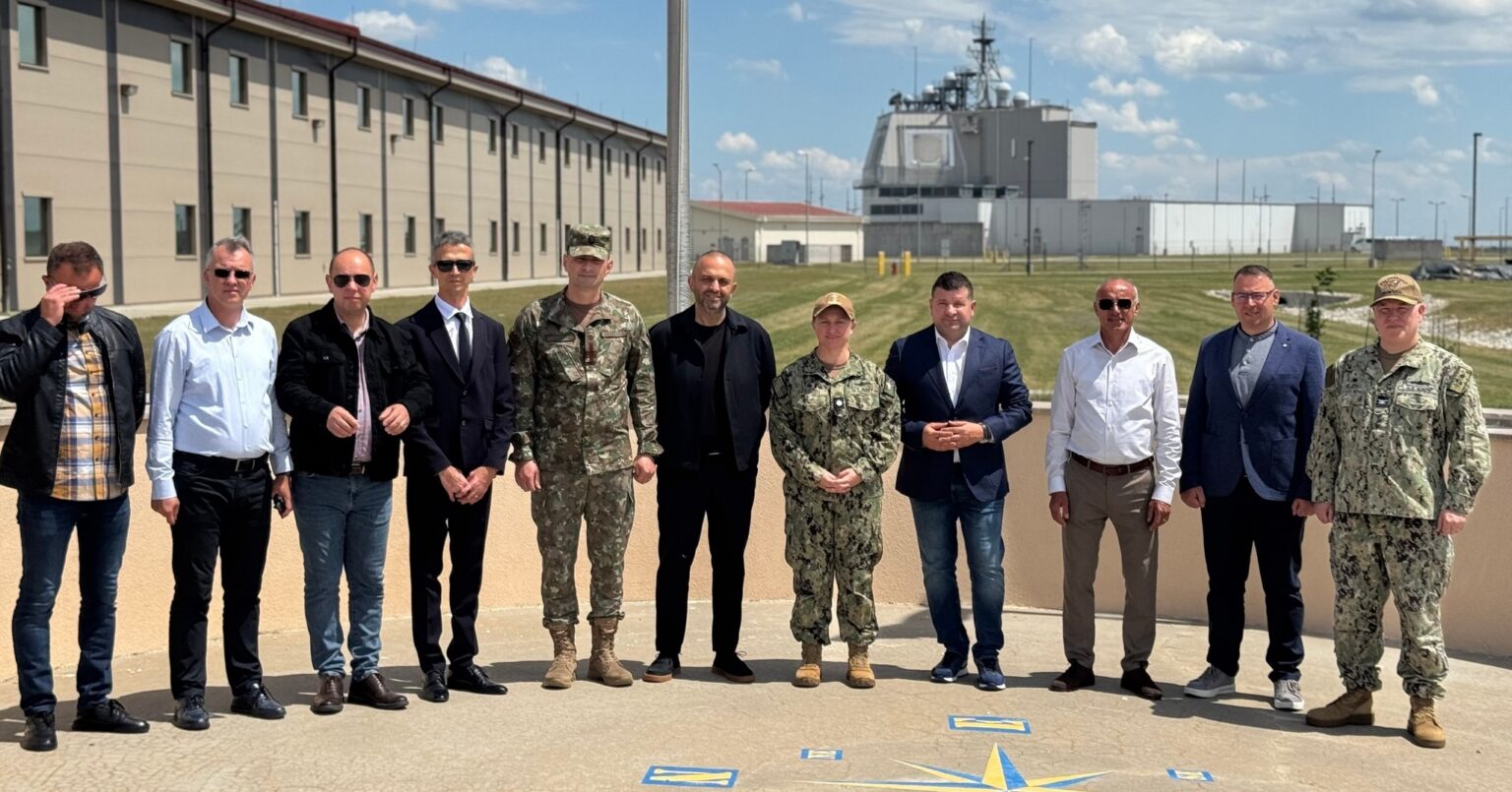Group of military personnel and civilians posing for a photo outside a base on a sunny day, with uniformed and civilian attire side by side.