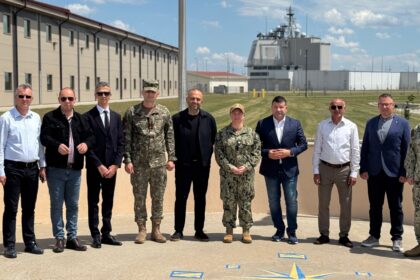 Group of military personnel and civilians posing for a photo outside a base on a sunny day, with uniformed and civilian attire side by side.