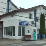 Hospital/clinic building with a blue sign for the emergency department; a medical staff member in teal scrubs stands by a small booth and bench near the entrance.
