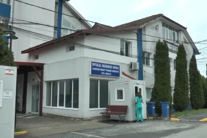 Hospital/clinic building with a blue sign for the emergency department; a medical staff member in teal scrubs stands by a small booth and bench near the entrance.