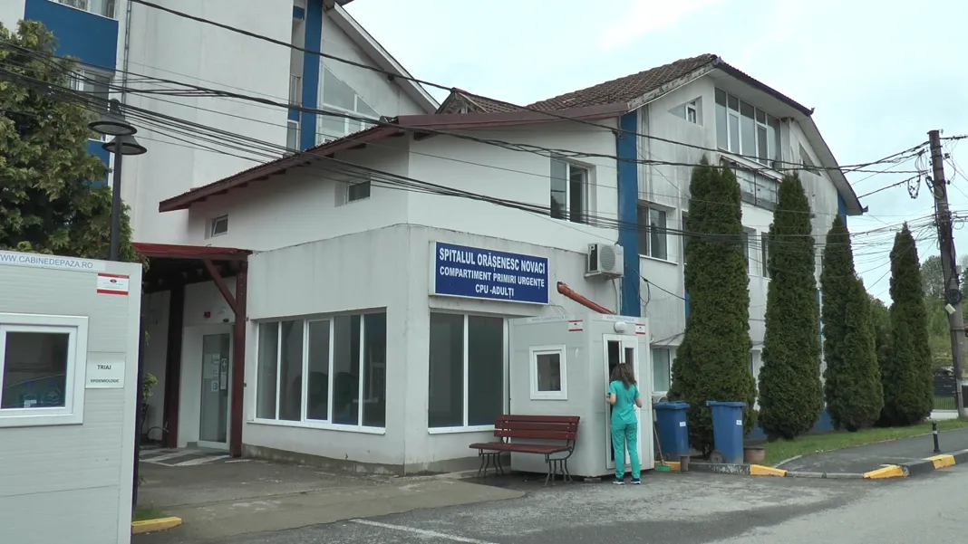 spital-novaci - Jurnalul Olteniei – Știri din Craiova și Oltenia Hospital/clinic building with a blue sign for the emergency department; a medical staff member in teal scrubs stands by a small booth and bench near the entrance.